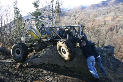 2003 Jeep TJ - Two individuals push a custom-built off-road buggy up a rocky, muddy slope in a mountainous, wooded area.