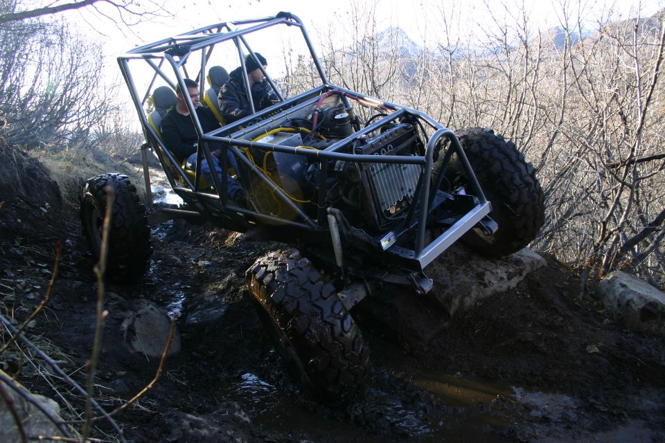 Off-road buggy with a roll cage, four people inside, navigating a muddy, rocky trail