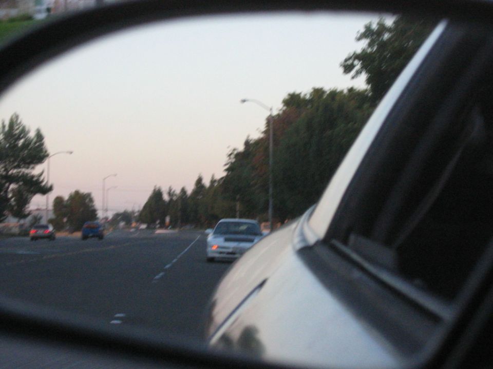 View from a car's side mirror shows traffic on a road at dusk with trees lining the side