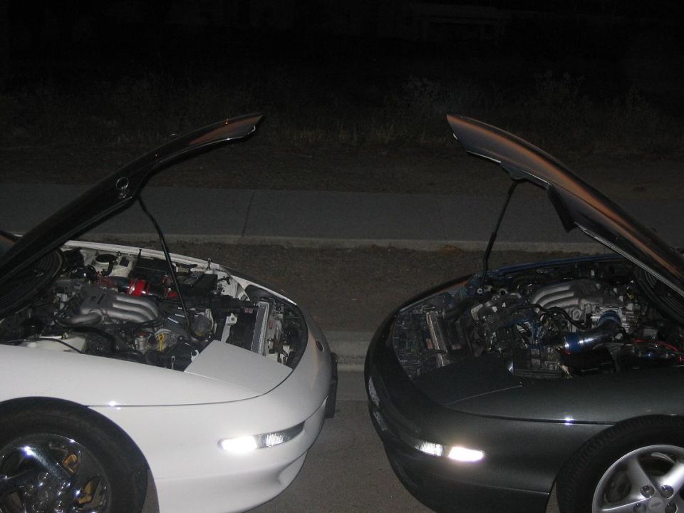 two cars, one white and one dark, parked side-by-side at night with hoods open showing engines