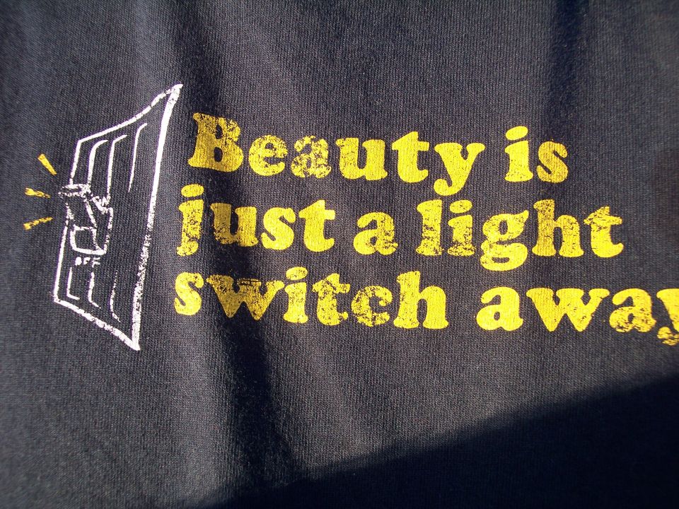 Black t-shirt with yellow text that reads "Beauty is just a light switch away" next to a graphic of a light switch