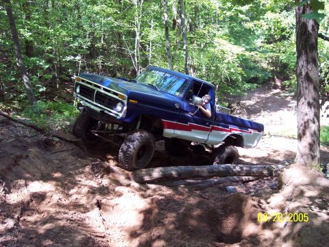 1977 Ford F-150 - A lifted blue pickup truck navigates a challenging dirt trail with logs and uneven terrain.