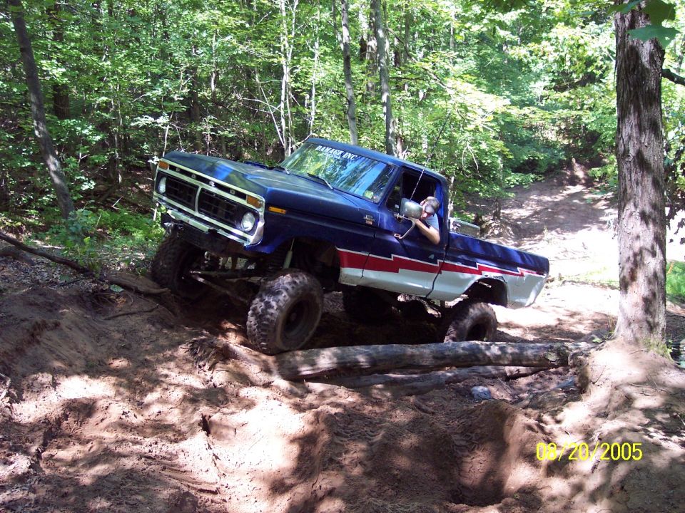 Blue pickup truck with red and white stripe on side, lifted with large tires, driving on a dirt trail through trees