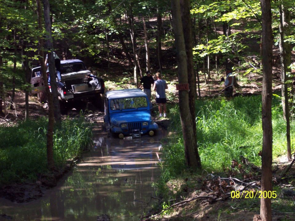 Blue Jeep Wrangler driving through a muddy stream in a forest, with another vehicle behind it