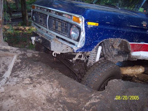 1977 Ford F-150 - A blue 1977 Ford F-150 is deeply stuck in mud, with its front tire submerged and suspension visible.