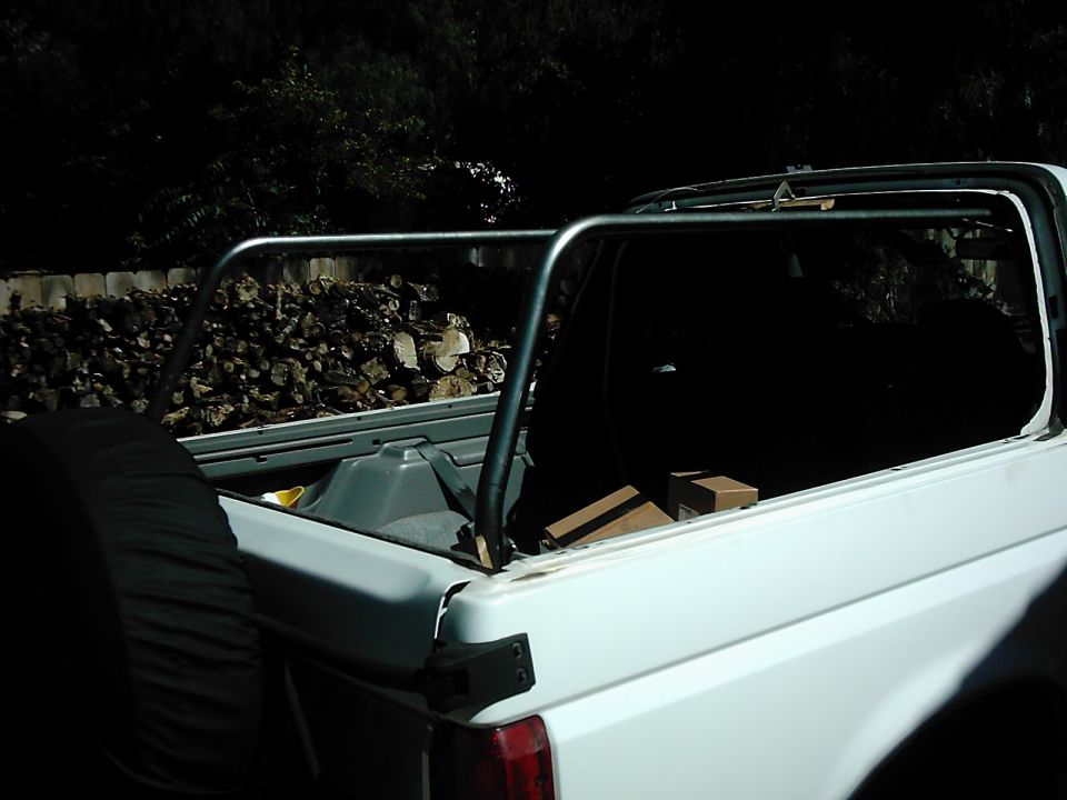 White SUV with roll cage, spare tire on the left, and firewood in the background. The vehicle appears to be a 1980s model