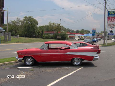 1978 Ford Bronco - A classic red Chevrolet is parked in a lot next to a newer car, with a gas station visible across the street.