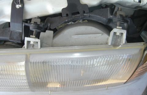 1991 Ford Taurus - Close-up under the open hood of a white car looking down at the top of a fluted rectangular composite headlight lens and its aluminum mount…