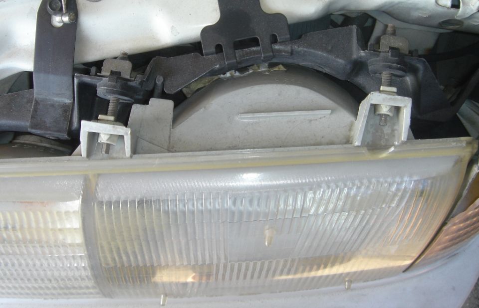 Close-up of headlight lens top and aluminum mounting bar under the hood of a white car