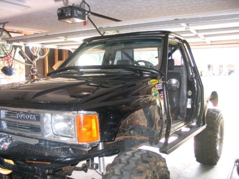 1986 Ford F-150 - This modified black Toyota truck features large off-road tires and a roll cage, seen from a 3/4 front angle in a garage.