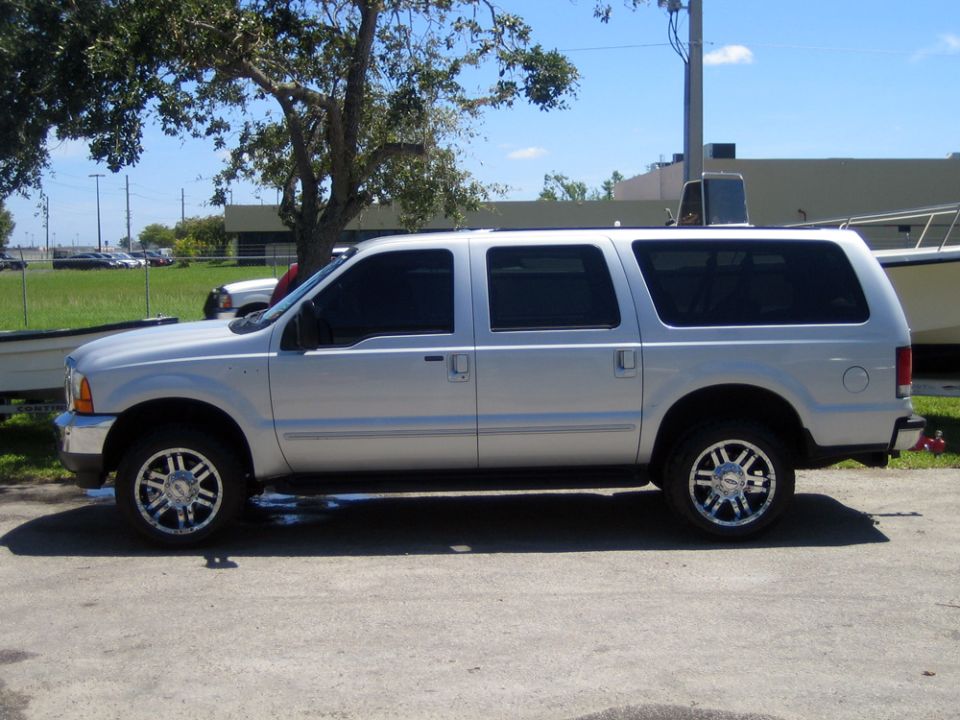 Silver Ford Excursion SUV, driver-side profile, chrome aftermarket wheels, parked outdoors near a tree