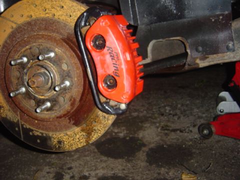 1998 Ford Taurus - A close-up view of an aftermarket racing brake caliper mounted on a rusty brake rotor.