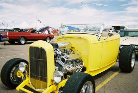 1972 Ford Ranchero - A yellow hot rod roadster is parked at an outdoor event, with a red pickup truck in the background.