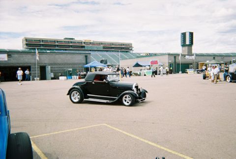 1972 Ford Ranchero - A black convertible hot rod is parked in front of a stadium with people milling around.