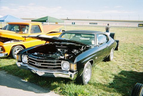 1972 Ford Ranchero - A black muscle car with its hood open is parked on grass next to an orange pickup truck.
