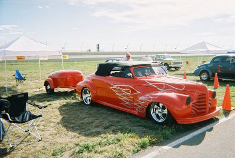 1972 Ford Ranchero - A bright red convertible with silver flame decals is parked next to a matching trailer, with other vehicles and tents in the background.