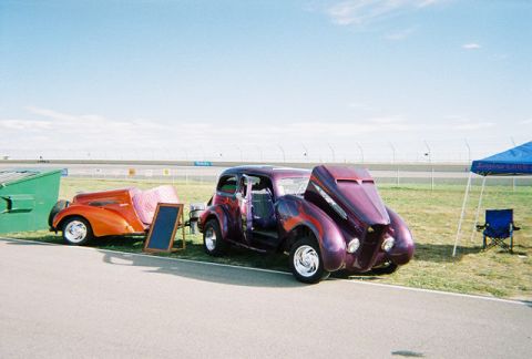 1972 Ford Ranchero - A purple hot rod with flames and an open hood sits beside an orange trailer at an outdoor event.
