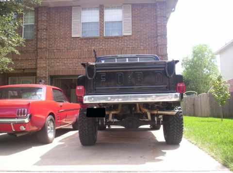 1984 Ford F-150 - A black Ford pickup truck with large tires is parked in front of a brick house, with a red Ford Mustang to its left.