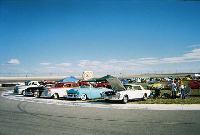 Several classic cars lined up on a track, some with hoods open. People are gathered around