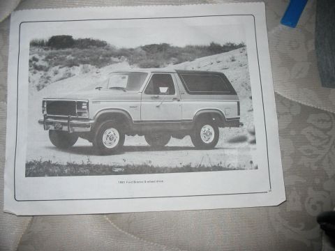 1986 Ford Bronco - A printed page lying on a patterned bedspread shows a black-and-white 3/4-front press photo of a two-tone full-size Bronco with a brush gua…