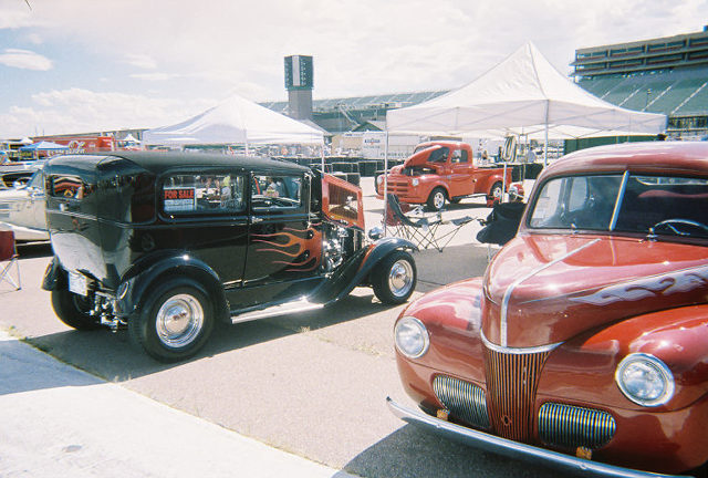 Black hot rod with flame decals and "FOR SALE" sign in window, parked next to a red car