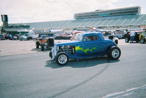 1972 Ford Ranchero - A blue hot rod with flame graphics is parked in front of a stadium, with other custom vehicles visible.