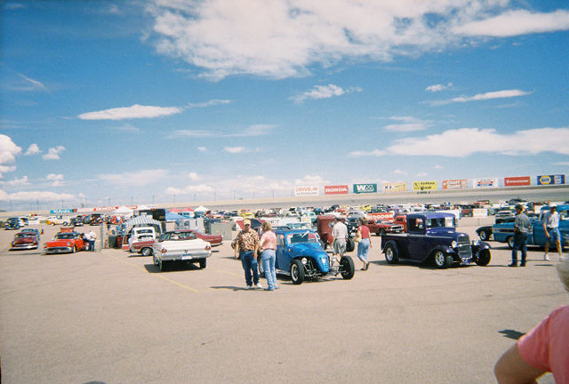 Outdoor car show with many classic cars and trucks parked on asphalt under a blue sky with clouds. People are milling around