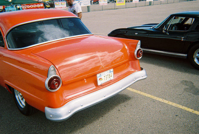 Orange 1955 Ford Fairlane rear bumper and taillights, black car in background