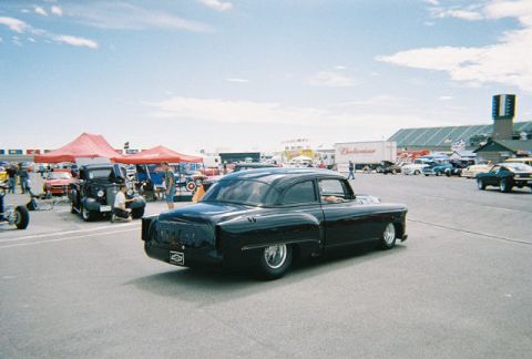 1972 Ford Ranchero - A black classic car with a lowered stance and chrome wheels is parked at an outdoor event with other vehicles and tents in the background.