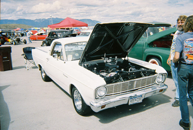 White Ford Ranchero with hood up showing engine, at a car show with mountains and onlookers