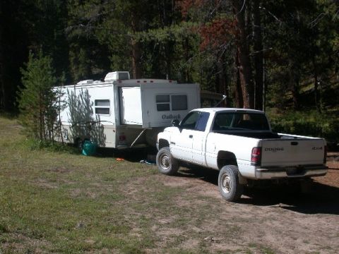 1994 Ford Bronco - A white Dodge pickup truck is hitched to a travel trailer, parked in a campsite surrounded by trees.