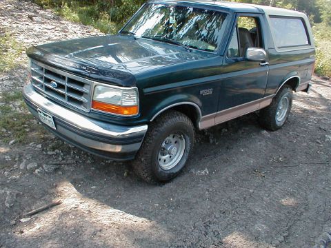 1994 Ford Bronco - A dark green 1994 Ford Bronco with tan trim is parked on a dirt path in a wooded area.