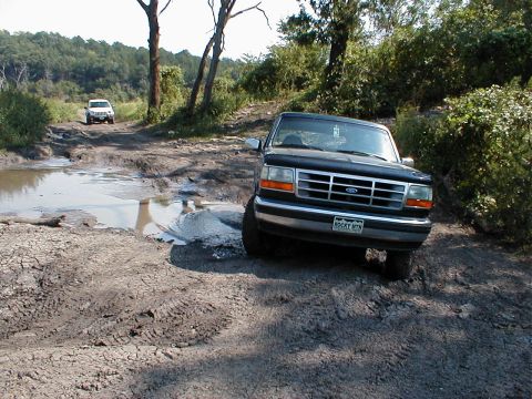 1994 Ford Bronco - A 1994 Ford Bronco navigates a muddy off-road trail with another vehicle in the distance.