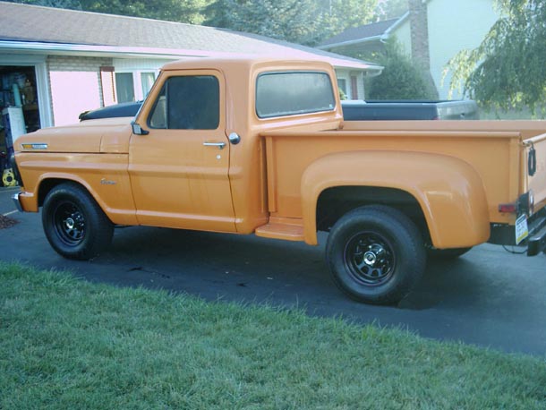 Orange short-bed stepside pickup parked on a lawn beside a brick house, black steel wheels