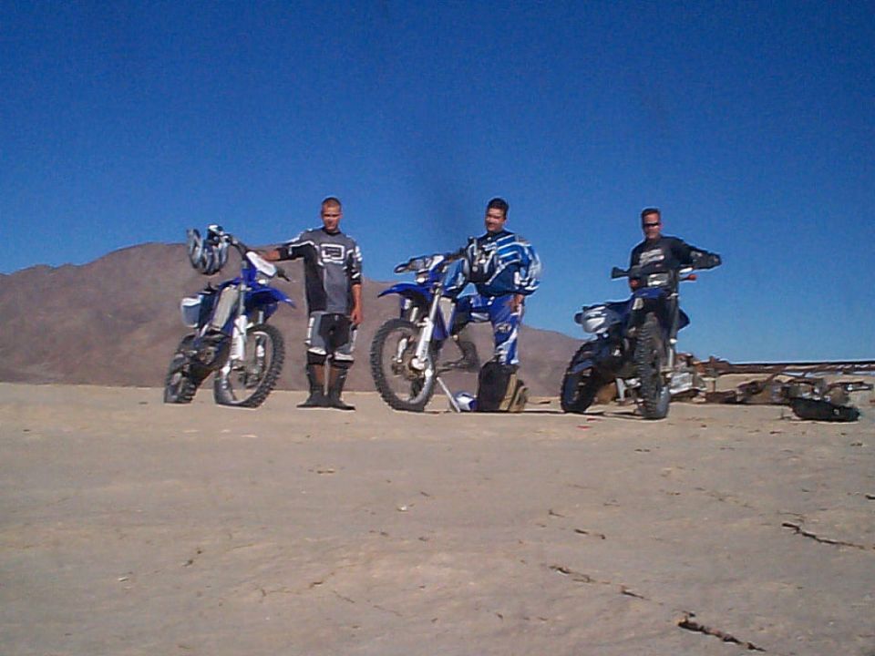 Three riders pose with blue Yamaha dirt bikes in desert with mountains behind