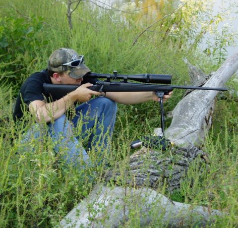 1995 Mercury Grand Marquis - Man aims a scoped rifle while kneeling in tall grass near a body of water.