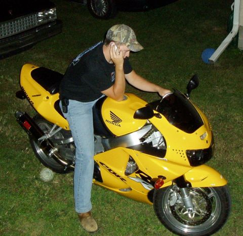 1995 Mercury Grand Marquis - A man sits on a bright yellow Honda motorcycle while talking on his cell phone.