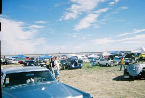 1972 Ford Ranchero - An outdoor car show under a blue sky with scattered clouds, numerous vehicles and tents.
