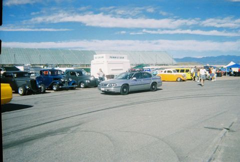 1972 Ford Ranchero - A silver police car is parked on asphalt with other vehicles and people in the background under a blue sky.