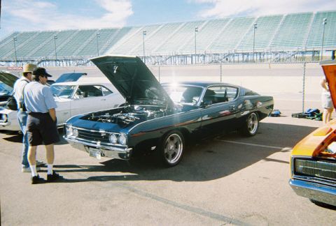 1972 Ford Ranchero - Three cars are parked at an event with grandstands in the background. The dark blue coupe has its hood open.