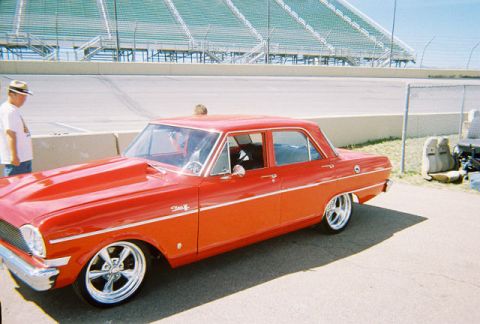 1972 Ford Ranchero - A red sedan with a prominent hood scoop and shiny chrome wheels is parked near a racetrack, with stadium seating visible in the background.