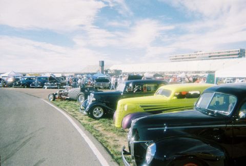 1972 Ford Ranchero - A line of vintage cars, including a black coupe and a bright yellow sedan, are parked on a sunny day with a stadium in the background.