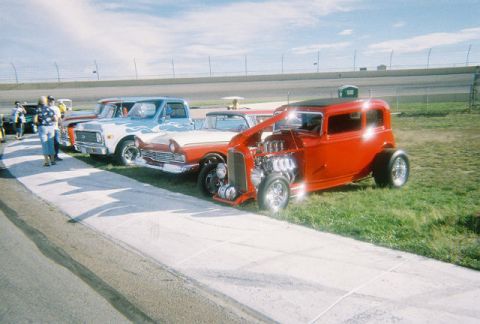 1972 Ford Ranchero - Several vehicles are parked on grass next to a racetrack, including a red hot rod with its hood up.