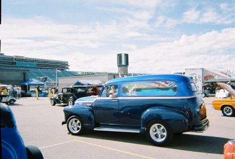 1972 Ford Ranchero - A blue panel van with a painted mural is parked at an outdoor event with other vehicles and grandstands visible.