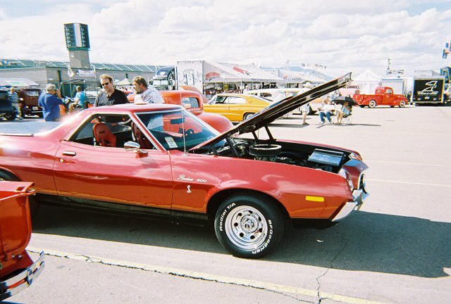 Red 1972 Ford Ranchero with hood open, parked on asphalt with other vehicles and tents in background