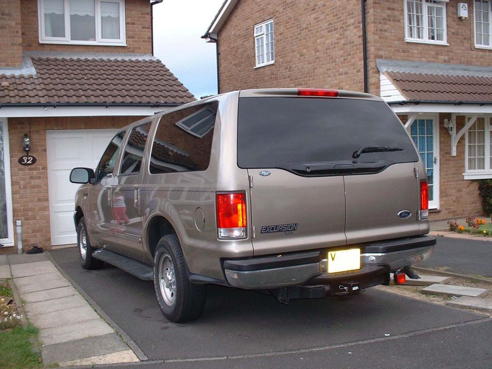 Rear three-quarter of gold Ford Excursion in brick-home driveway with tow hitch