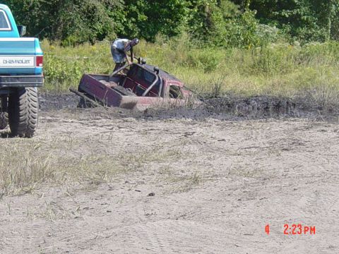 1995 Dodge 1500 - A red pickup truck is stuck in the mud while a person attempts to free it. Another blue truck is partially visible on the left.