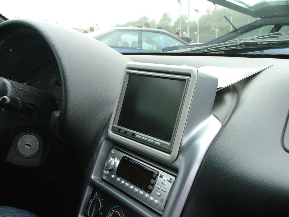 Car dashboard showing an aftermarket screen and stereo unit. Another car is visible through the windshield
