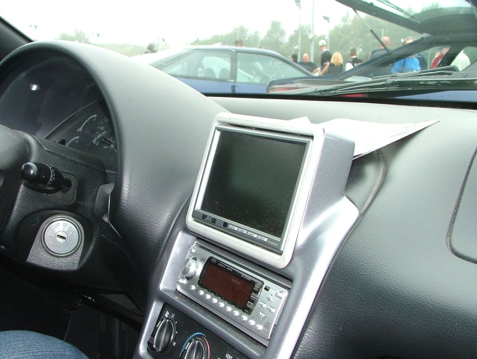 Car interior showing dashboard, steering wheel, and aftermarket stereo system. Another car is visible through the windshield