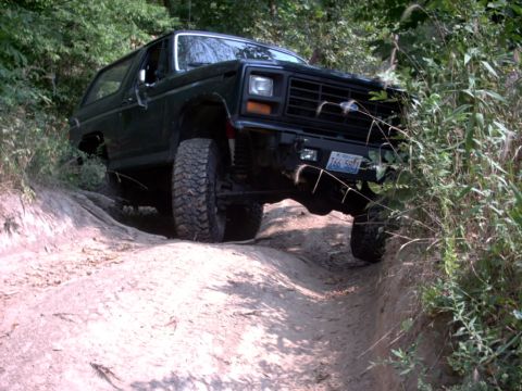 1986 Ford Bronco - A black Ford Bronco navigates a challenging dirt path surrounded by dense foliage.